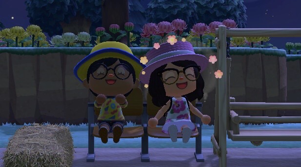 A screenshot of me and Twigs in Animal Crossing. We're wearing matching outfirs but in different colors. I'm wearing a yellow bucket hat with a blue rim, round glasses, a retro shirt, and jeans. She's wearing a pink bucket hat, geometric glasses, and a dress with flowers on it. We both are doing happy emotes, and there are flowers surrounding Twigs.